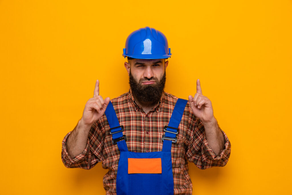 bearded builder man construction uniform safety helmet looking with serious face pointing with index fingers up scaled
